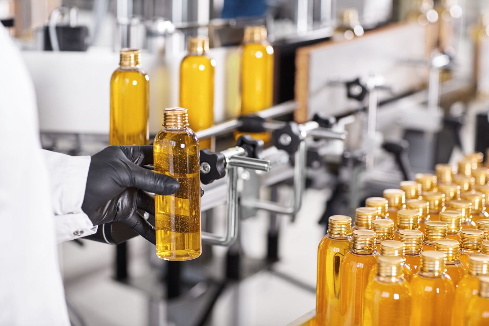 Process of making new healthcare products on big modern factory.Scientists wearing white medical gown and rubber black gloves holding glass bottle with yellow thick liquid going to put it on conveyor